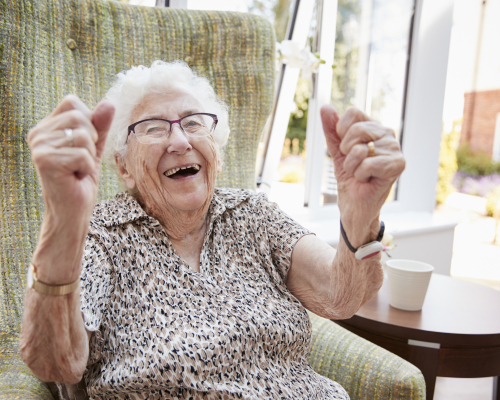 Portrait Of Excited Senior Woman Sitting In Chair In Lounge Of Retirement Home