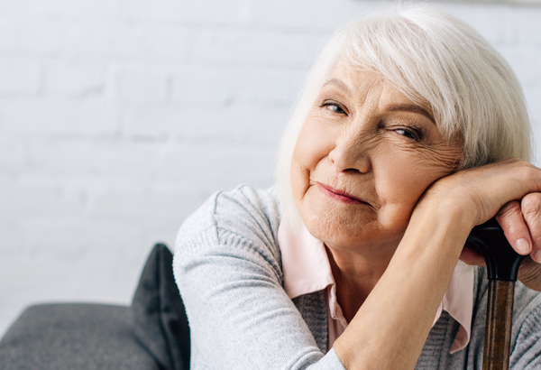 panoramic shot of smiling senior woman with wooden cane looking at camera in apartment